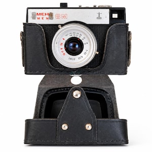 Puede incluir: Una cámara vintage MEH 8M con cuerpo plateado y negro y un objetivo circular prominente. La cámara está alojada en una funda protectora de cuero negro con una solapa frontal abatible asegurada con tachuelas metálicas visibles.
