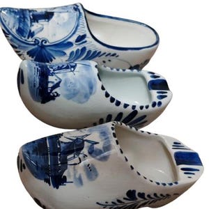 May include: Three white ceramic clogs with blue floral and landscape designs. The clogs are stacked, showcasing the traditional Dutch style. Each clog features a rounded toe and a hollow interior.