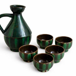 Puede incluir: Un juego de sake de cerámica verde con una jarra a juego y cinco tazas pequeñas. La jarra tiene un asa y una tapa. Las tazas y la jarra están decoradas con rayas verdes verticales. El interior de las tazas es marrón.