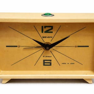 Pode incluir: Despertador vintage creme com mostrador retangular e design retrô. O mostrador do relógio é amarelo claro com algarismos e ponteiros pretos. O relógio tem o texto "SEVANI" e "MADE IN USSR". Um botão verde está no topo.