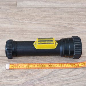 Könnte beinhalten: Eine schwarze zylindrische Taschenlampe mit einem gelben rechteckigen Schaltergehäuse, das die Aufschrift "MAGNETIC FLASHLIGHT" trägt. Die Taschenlampe liegt über einem gelben Maßband, das eine Länge von etwa 21,5 cm anzeigt.