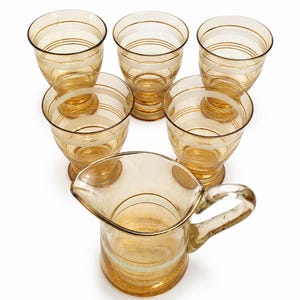 Könnte beinhalten: Ein Set aus fünf bernsteinfarbenen Gläsern und einem passenden Krug. Jedes Teil ist mit einer Reihe horizontaler weißer und goldfarbener Bänder verziert, die den Körper umschließen. Der Krug verfügt über einen geschwungenen Henkel.