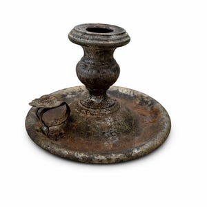 May include: An antique metal candlestick with a circular base and central stem. The candlestick has a weathered, aged appearance with a dark, mottled finish. A small, curved piece is attached to the base.