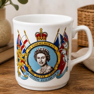 Könnte beinhalten: Ein weißer Keramikbecher mit einem Gedenkmotiv zum silbernen Thronjubiläum von Königin Elizabeth II. Das Design zeigt ein Porträt der Königin, eine Krone, Flaggen und den Schriftzug TO COMMEMORATE THE SILVER JUBILEE OF QUEEN ELIZABETH II 1952-1977.