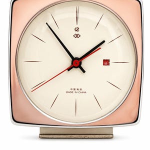 Può includere: Una sveglia quadrata, color oro rosa, con quadrante color crema. L'orologio presenta lancette nere, una lancetta dei secondi rossa e un datario. Il quadrante include la scritta "MADE IN CHINA" e caratteri cinesi. L'orologio poggia su una base chiara.