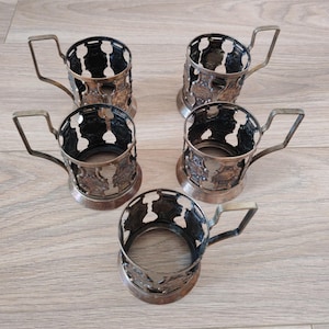 Peut inclure: Un ensemble de cinq porte-gobelets métalliques, chacun présentant un design ajouré avec un motif répétitif et une poignée unique. Les supports ont une finition sombre et vieillie ainsi qu'une forme cylindrique pour maintenir un verre.