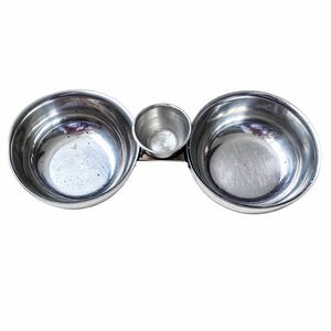 Peut inclure: Un plat de service en acier inoxydable à trois compartiments. Deux grands bols encadrent un petit bol central, le tout relié par un support métallique. Les bols ont une surface polie et réfléchissante, idéale pour servir des collations ou des sauces.