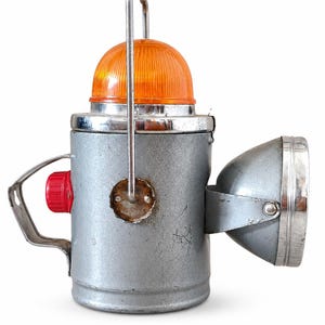 May include: A vintage silver-toned metal lantern featuring a cylindrical body, a bright orange ribbed dome top, and a side-mounted spotlight. It includes a metal carrying handle, a red circular button on the side, and a metallic finish with wear.