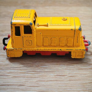 Puede incluir: Un tren de juguete amarillo con una base roja. El tren de juguete tiene detalles en negro y el número "5" en el lateral. El tren de juguete está sobre una superficie de madera.