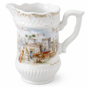 Może przedstawiać: Biały dzbanek z porcelany z falistym brzegiem ze złotymi akcentami oraz uchwytem. Korpus zdobi kolorowa ilustracja sceny wystawowej z budynkami, palmami oraz napisem "Indische Gewerbe" i "Ausstellung 1886".