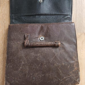 Op de afbeelding: Een donkerbruine lederen portfolio met een getextureerd oppervlak en een horizontaal handvat aan de voorzijde. De portfolio heeft een zwarte lederen binnenflap met een zilverkleurige metalen drukknoop in het midden van het voorpaneel.