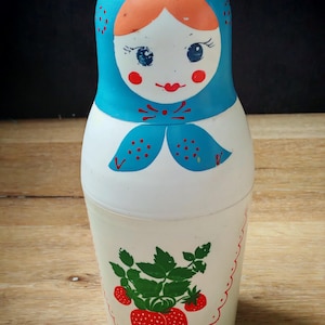 May include: A traditional Russian Matryoshka doll. The doll has a blue headscarf and a white body. The face is painted with rosy cheeks and a red mouth. The body is decorated with a strawberry design, green leaves, and a scalloped border.