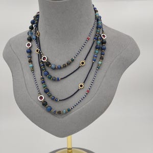 Op de afbeelding: Een meerstrengs ketting met donkerblauwe kraaltjes afgewisseld met grotere ronde blauwe en groene stenen kralen. Het ontwerp bevat kleine goudkleurige metalen tussenstukjes en ronde accenten met ingebedde rode en zilverkleurige centra.