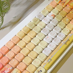 May include: A mechanical keyboard featuring a white frame and a gradient colour scheme of orange, yellow, and cream keycaps. Several keys display custom illustrations of landscapes and small icons. The spacebar features a printed scene of a road.