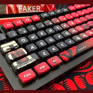Könnte beinhalten: Eine mechanische Tastatur in Schwarz und Rot mit individuellen Tastenkappen. Die Tasten sind eine Mischung aus Schwarz und Rot, einige mit Grafiken und dem Namen "FAKER". Die Tastatur liegt auf einer rot-schwarz gemusterten Oberfläche.