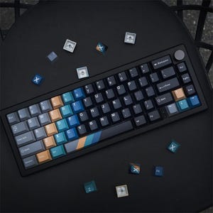 May include: A black mechanical keyboard with colorful keycaps. The keycaps feature a mix of blue, orange, and gray colors. Several loose keycaps with an "X" design are scattered around the keyboard on a black surface.
