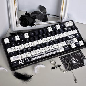 Może przedstawiać: Czarno-biała klawiatura mechaniczna z niestandardowymi nakładkami na klawisze. Nakładki na klawisze mają różne wzory, w tym motyle, wzory w szachownicę i tekst. Czarna róża i ozdobna karta znajdują się w tle. Brelok w kształcie nietoperza i pióra są również obecne.