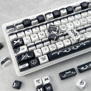 May include: A white mechanical keyboard with black and white keycaps. The keycaps feature various designs, including bows, hearts, and polka dots. Some keys are black with white text, while others are white with black text. The keyboard has a sleek, modern design.