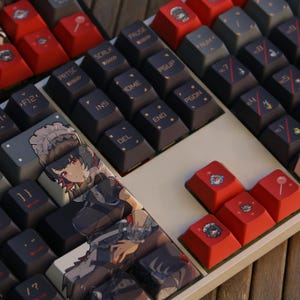 Może przedstawiać: Zbliżenie klawiatury mechanicznej z niestandardowymi nakładkami na klawisze. Nakładki na klawisze są mieszanką czerni, czerwieni i szarości. Niektóre nakładki na klawisze zawierają ilustracje w stylu anime. Klawiatura ma białą podstawę i jest ustawiona na drewnianej powierzchni.