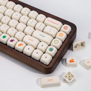 以下が含まれることがあります： 木製のフレームとオフホワイトのキーキャップを備えたキーボード。キーには、オレンジ、緑、紫などの色でカラフルなフルーツアイコンとテキストが表示されています。レトロな美学のキーボードです。