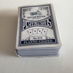Könnte beinhalten: Ein Kartenspiel mit weißem und blauem Design. Die Kartenbox trägt die Aufschrift "MOTOR BRAND HIGH QUALITY PLAYING CARDS" und "No. 976 PLASTIC COATED". Das Spiel ist auf einer weißen Oberfläche gestapelt.