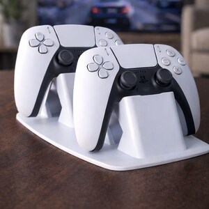 Könnte beinhalten: Zwei weiße und schwarze Gamecontroller auf einem weißen Ständer. Die Controller haben ein symmetrisches Design mit verschiedenen Tasten und Richtungspads. Der Ständer ist rechteckig und abgewinkelt, um die Controller aufrecht zu halten. Im Hintergrund ist ein Fernseher zu sehen.