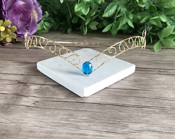Puede incluir: Una diadema de alambre dorado con una piedra preciosa azul en el centro. La diadema está decorada con delicados remolinos de alambre.