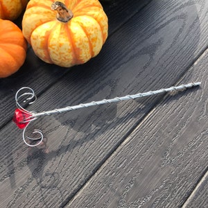 Puede incluir: Una varita de plata con una gema roja en la parte superior y un diseño decorativo de espiral. La varita está sobre una superficie de madera oscura.