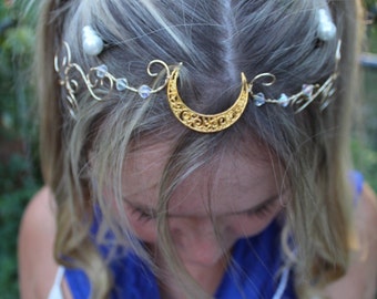 Puede incluir: Una diadema dorada con forma de media luna y vid con cuentas transparentes y perladas. La diadema se lleva sobre cabello rubio, con un atuendo de estilo marinero azul, blanco y rojo visible debajo. El atuendo tiene un gran lazo rojo.