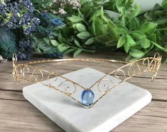 Puede incluir: Una corona de alambre dorado con un diseño delicado y en espiral y una sola piedra preciosa azul en el centro.