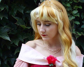 May include: A woman wearing a pink and red dress with a silver crown and necklace. She has long blonde hair with a light blonde streak. She is wearing a red rose brooch.