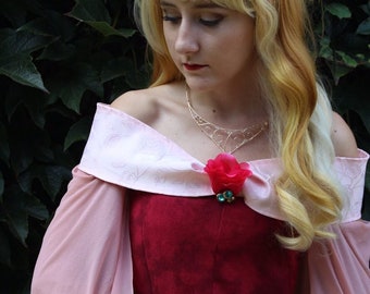 Könnte beinhalten: Eine Frau in einem rosa und roten Kleid mit einer goldenen Krone und einer roten Rosenbrosche. Sie hat langes blondes Haar und schaut nach unten.