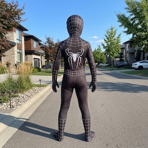 May include: Black Spider-Man costume with a white spider emblem on the back. The full-body suit features a web pattern and includes a hood, gloves, and foot coverings. The costume is worn outdoors on a paved road.