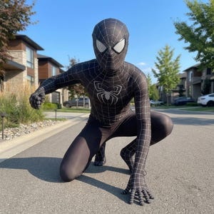 May include: A black Spider-Man costume with a white spider emblem on the chest and white web detailing. The full-body suit includes a mask with white eye lenses. The person is crouching on a paved road.