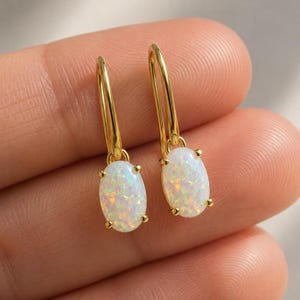 May include: A pair of gold-colored drop earrings. Each earring features an oval, iridescent white opal stone suspended from a slender, curved gold-tone hook. The opals are secured by four small gold prongs.