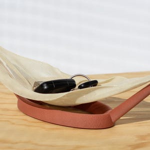 Pode incluir: Uma moderna e minimalista bandeja multiuso em terracota quente. A bandeja apresenta uma rede de malha bege suspensa entre dois suportes curvos. Dois objetos pretos e um anel prateado repousam dentro.