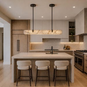 May include: A rectangular linear pendant light fixture with a translucent, veined stone shade suspended by two thin black rods above a kitchen island. Three light-colored upholstered stools with dark wooden legs are positioned at the island.