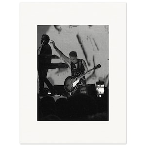 Può includere: Fotografia in bianco e nero di un'esibizione musicale dal vivo. Una persona suona una chitarra elettrica, mentre un'altra suona la tastiera sullo sfondo. Il pubblico è visibile in primo piano, con un grande schermo dietro gli artisti.