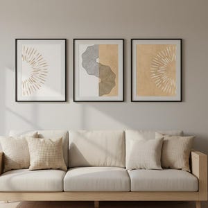 May include: Three framed abstract art prints with black frames hang above a beige sofa with matching pillows. The art features geometric designs in neutral tones. A small side table and vase complete the room's decor.