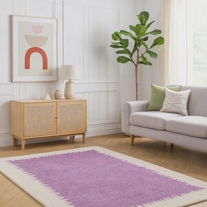 Puede incluir: Una alfombra rectangular con un centro lila y un borde crema. La alfombra tiene un aspecto texturizado y está colocada sobre un suelo de madera. La habitación también cuenta con un sofá de color claro, un mueble de madera y una planta grande.