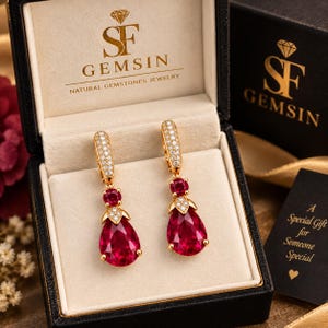 May include: A pair of gold drop earrings featuring pear-shaped red gemstones, small round red stones, and pavé-set white diamonds. The earrings are displayed in a black jewellery box with the text SF GEMSIN NATURAL GEMSTONES JEWELRY.