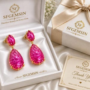 May include: A pair of gold-tone drop earrings featuring vibrant pink faceted stones in oval and teardrop shapes. The earrings are presented in a white satin-lined box labelled SFGEMSIN with a matching gift box and a thank you card included.