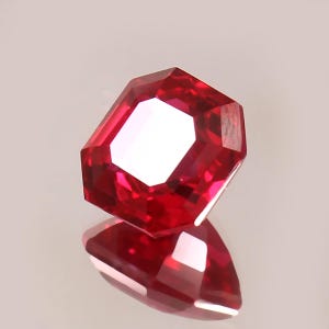May include: A faceted, octagonal-cut ruby gemstone. The gem is a deep, vibrant red with a reflective surface, showcasing its internal structure and brilliance. The gem is displayed on a reflective surface, creating a mirror image.