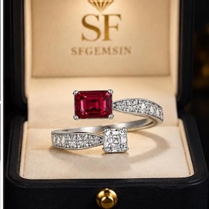 Puede incluir: Un anillo abierto con una piedra roja de corte esmeralda rectangular y una piedra blanca de corte cuadrado, sobre una banda plateada con pequeñas piedras blancas. El anillo se presenta en una caja negra con el texto SF SFGEMSIN.