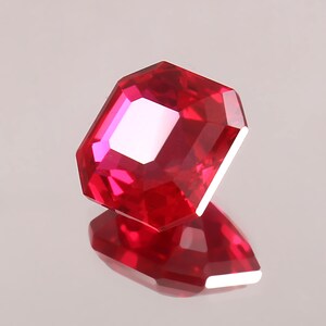 以下が含まれることがあります： ファセットカットされた長方形の赤い宝石。透明度が高く、非常に反射性が高く、明るく輝いています。反射する表面に置かれており、鏡像を作り出しています。