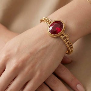 May include: A gold-toned metal bracelet featuring a large, oval-shaped, deep red faceted gemstone set in a rope-textured frame. The band consists of a thick chain-link design and includes a small crown emblem detail near the central stone setting.