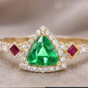 May include: A gold ring featuring a central trillion-cut green gemstone surrounded by a halo of small white diamonds. Two square-cut red rubies are set on either side of the center stone, with additional small white diamonds along the band.