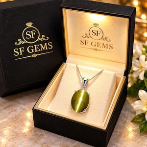 Puede incluir: Un colgante de piedra ojo de gato de color verde oliva con forma ovalada en una cadena de plata, presentado dentro de un joyero negro de la marca SF GEMS con luz interior. La tapa presenta el logotipo de SF GEMS en oro, a juego con el interior.