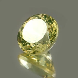 May include: A faceted, oval-shaped gemstone with a pale yellow hue. The gem is highly reflective, with a bright, white center and a complex pattern of internal reflections. The stone is set against a neutral background, with its reflection visible below.