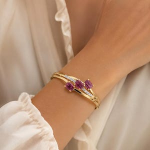 May include: A gold cuff bracelet featuring three emerald-cut magenta gemstones arranged in a row, accented by a thin diagonal line of small clear pavé-set stones that cross over the band, creating a layered and polished metallic appearance.